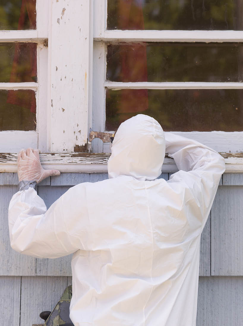 A house painter in a hazmat suit scrapes off dangerous lead paint from a window sill 