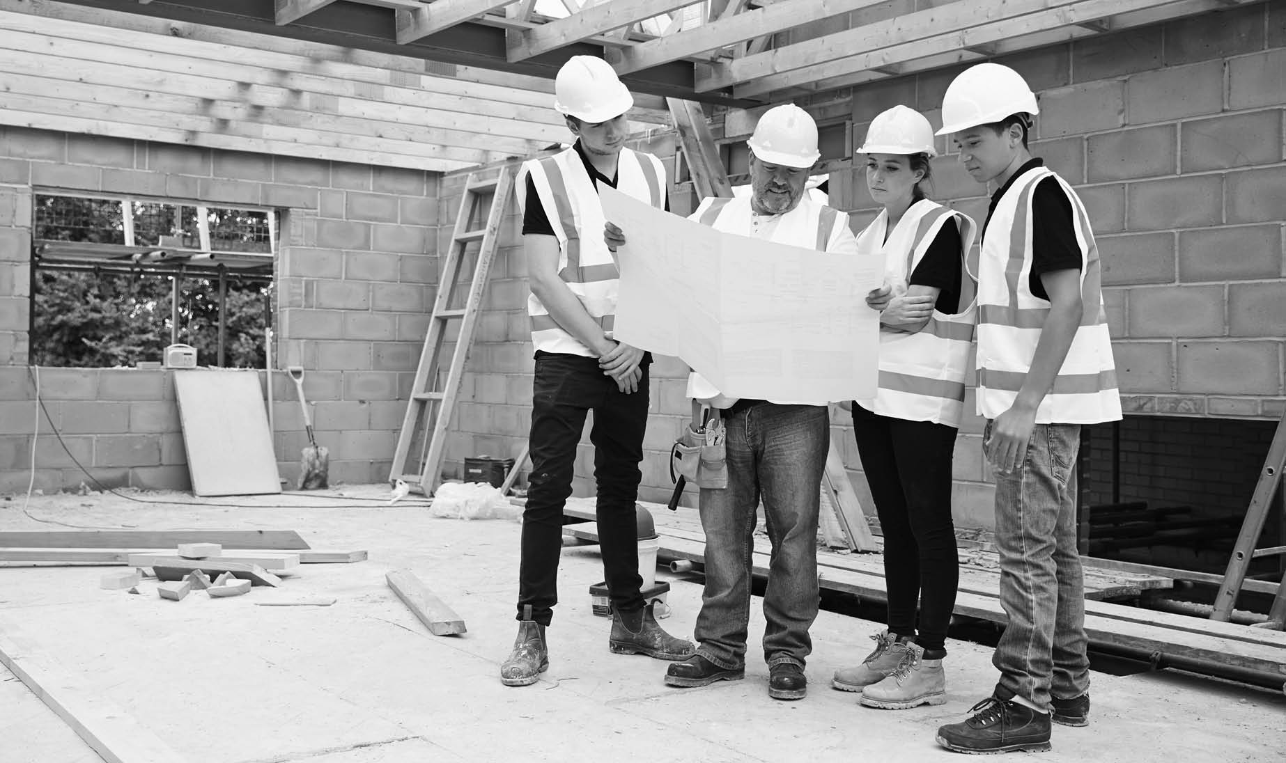 Builder On Building Site Looking At Plans With Apprentices