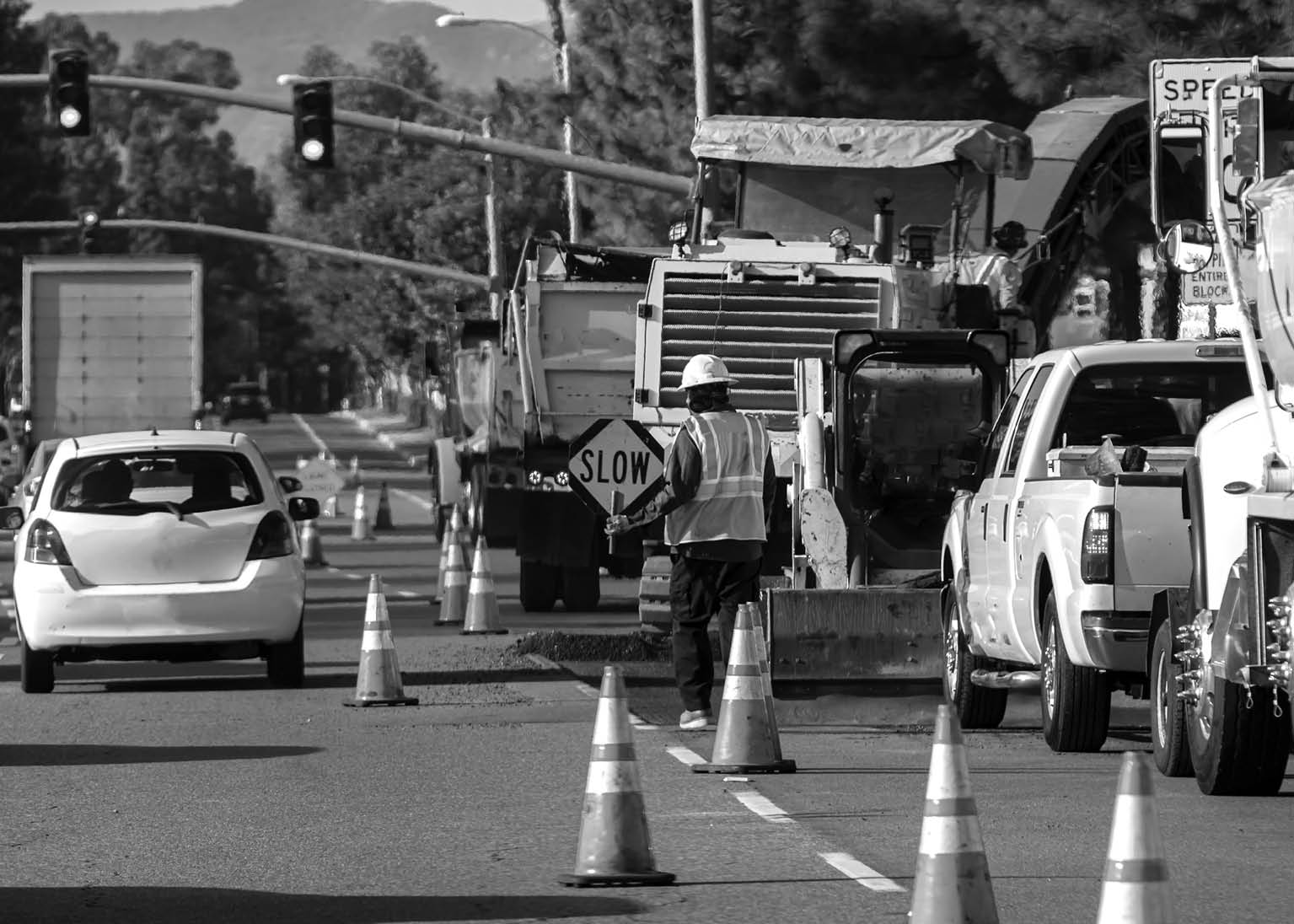 Wide view of raod construction site and a nan holding traffic sign that says SLOW  Bright orange traffic cones and trucks and cars are visible