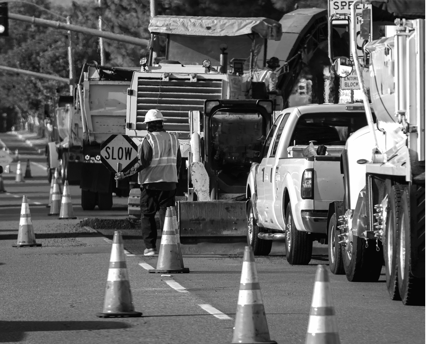 Wide view of raod construction site and a nan holding traffic sign that says SLOW  Bright orange traffic cones and trucks and cars are visible