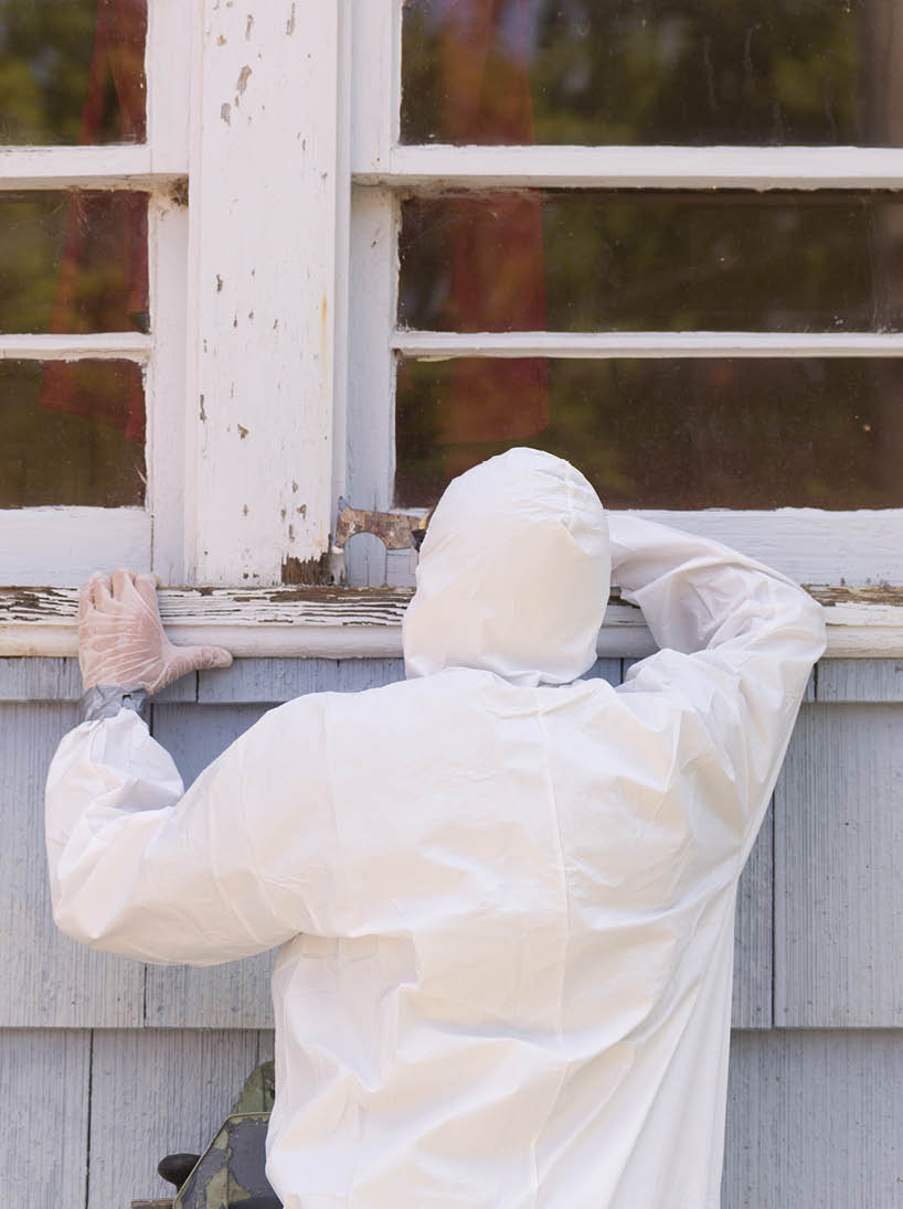 A house painter in a hazmat suit scrapes off dangerous lead paint from a window sill 